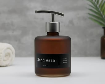 Puede incluir: Un dispensador de jabón de manos de vidrio esmerilado ámbar de forma cuadrada con una bomba de metal plateado. La etiqueta negra presenta el texto blanco Hand Wash, 250ml, y una lista de tres elementos: refill, reuse, repeat, separados por una línea.