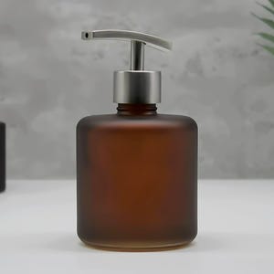 May include: A cylindrical soap dispenser made of frosted amber glass with a brushed silver metal pump mechanism. The bottle has a flat base and a smooth, matte finish, featuring a curved spout on the pump head for dispensing liquid soap or lotion.