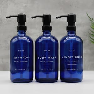 Könnte beinhalten: Drei dunkelblaue Glasflaschen mit schwarzen Pumpen, beschriftet mit "SHAMPOO", "DUSCHGEL" und "SPÜLUNG". Jede Flasche hat weißen Text mit der Aufschrift "NATÜRLICHE INHALTSSTOFFE" und "500ml | 16.91 fl oz". Die Flaschen stehen auf einer weißen Oberfläche.