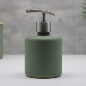 May include: A cylindrical soap dispenser with a matte sage green finish and a brushed silver metal pump mechanism. The container features a smooth, opaque surface and a flat base, designed for dispensing liquid soap or lotion in a bathroom or kitchen.