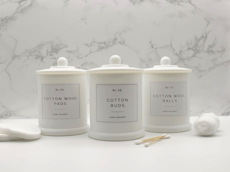 Może przedstawiać: Trzy białe ceramiczne słoiki z pokrywkami, każdy oznaczony innym rodzajem produktu bawełnianego: "Cotton Wool Pads", "Cotton Buds" i "Cotton Wool Balls". Każdy słoik ma czarną etykietę z nazwą produktu i nadrukiem "100% Organic".