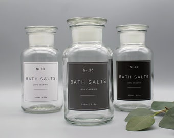 Può includere: Tre barattoli di vetro trasparente con etichette nere che recitano "No. 30 Bath Salts 100% Organic 500ml | 420g". I barattoli sono su una superficie grigia con foglie verdi.