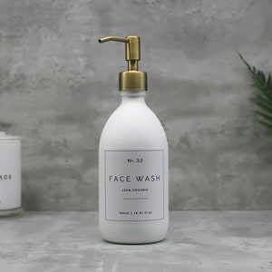 May include: White face wash dispenser with a gold-colored pump. The label reads "FACE WASH" and "100% ORGANIC". The bottle is 500ml / 16.91 fl oz. A container of cotton pads is in the background.