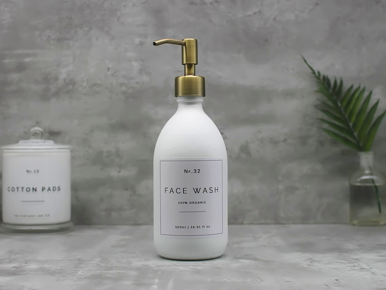 May include: White face wash dispenser with a gold-colored pump. The label reads "FACE WASH" and "100% ORGANIC". The bottle is 500ml / 16.91 fl oz. A container of cotton pads is in the background.