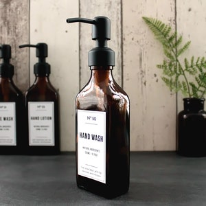 May include: Three amber glass bottles with black pump tops and white labels. The labels read "Face Wash", "Hand Lotion", and "Hand Wash". The bottles are on a dark grey surface.