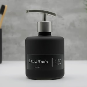 May include: A black hand wash dispenser with a silver pump. The label reads "Hand Wash" and "250ml". The dispenser is part of a bathroom set, with a matching black toothbrush holder and toothbrushes visible in the background.