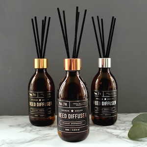 Op de afbeelding: Drie bruine glazen rietdiffusers met zwarte rietjes. De diffusers hebben gouden en zilveren doppen en labels met de tekst "No. 71 Reed Diffuser" en "No. 74 Reed Diffuser".