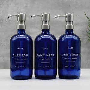 May include: Three dark blue glass bottles with silver pump dispensers. Each bottle is labelled with white text: Shampoo, Body Wash, and Conditioner. The bottles are 500ml / 16.91 fl oz.