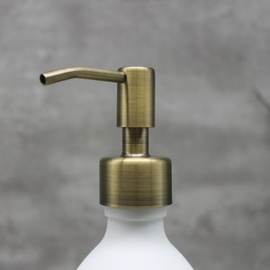 May include: Close-up of a white soap dispenser with a brass-colored pump. The pump has a curved spout and a cylindrical base. The dispenser is against a gray background.