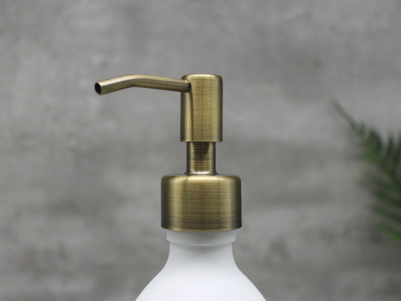 May include: Close-up of a white soap dispenser with a brass-colored pump. The pump has a curved spout and a cylindrical base. The dispenser is against a gray background.