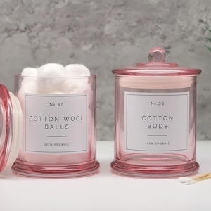 Op de afbeelding: Twee roze glazen apothekerspotten met deksels, met de labels "Cotton Wool Balls" en "Cotton Buds". Eén pot is open en gevuld met witte wattenbolletjes. De andere pot is gesloten. De labels luiden "100% Organic".