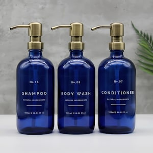 May include: Three dark blue glass bottles with bronze-coloured pump dispensers. Each bottle is labelled with white text: "SHAMPOO", "BODY WASH", and "CONDITIONER". The bottles are 500ml / 16.91 fl oz.