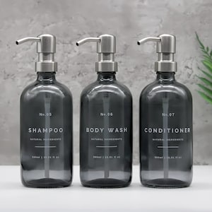 May include: Three grey glass pump bottles labelled "SHAMPOO", "BODY WASH", and "CONDITIONER". Each bottle has a silver pump and holds 500ml (16.91 fl oz) of product. The bottles are arranged on a white surface.