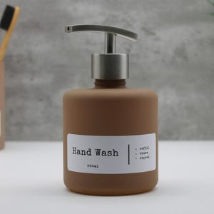 Puede incluir: Un dispensador de jabón de manos marrón con una bomba plateada y una etiqueta blanca que dice "Hand Wash" y "250ml". La etiqueta también incluye las palabras "refill, reuse, repeat". Un porta cepillos de dientes es visible en el fondo.