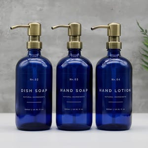 May include: Three dark blue glass bottles with brass-colored pumps. The bottles are labeled "Dish Soap", "Hand Soap", and "Hand Lotion". Each bottle holds 500ml or 16.01 fl oz of product. The labels also state "Natural Ingredients".