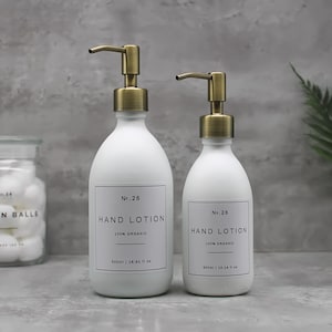 May include: Two white hand lotion dispensers with brass-colored pumps. The bottles have labels that read "Hand Lotion 100% Organic". The larger bottle holds 16.91 fl oz. A glass jar of cotton balls is in the background.