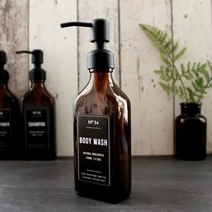 Puede incluir: Tres botellas de vidrio ámbar con tapas de bomba negras y etiquetas. Las botellas están etiquetadas como "Acondicionador", "Champú" y "Gel de ducha". Las botellas están sobre una superficie negra con una planta de helecho verde en un jarrón de vidrio marrón.