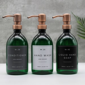 Op de afbeelding: Drie groene glazen flessen met koperen pompen en zwarte etiketten. De etiketten hebben witte tekst die "No. 29 Conditioner 100% Organic 500ml | 16.91 fl oz", "No. 25 Hand Wash 100% Organic 500ml | 16.93 fl oz" en "No. 42 Liquid Hand Soap 500ml | 16.91 fl oz" luidt.