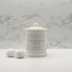Może przedstawiać: Biały ceramiczny słoik z pokrywką, oznaczony etykietą "No. 37 Cotton Wool Balls 100% Organic". Dwie białe kulki z waty znajdują się przed słoikiem na białym marmurowym blacie.