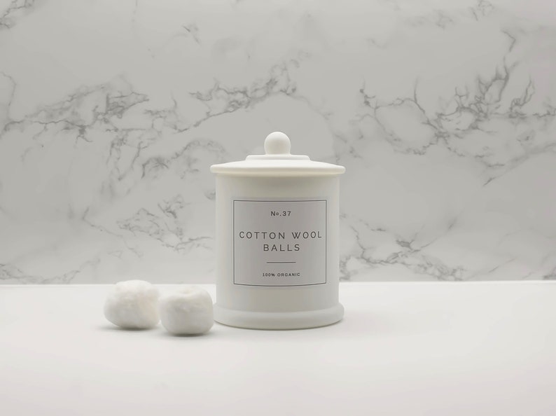 Może przedstawiać: Biały ceramiczny słoik z pokrywką, oznaczony etykietą "No. 37 Cotton Wool Balls 100% Organic". Dwie białe kulki z waty znajdują się przed słoikiem na białym marmurowym blacie.