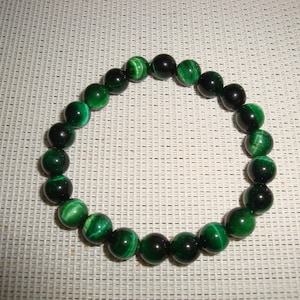 Puede incluir: Un brazalete de cuentas de ojo de tigre verde con rayas verde oscuro y negras. El brazalete está hecho de cuentas redondas y está enhebrado en un cordón elástico transparente.