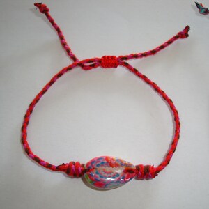 Op de afbeelding: Een rood, bruin en roze gevlochten armband met een kleurrijke schelpbedel.