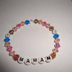 Op de afbeelding: Een kralenarmband met verschillende kleuren, waaronder roze, blauw en bruin. De armband heeft het woord "MAMAN" in witte letters.