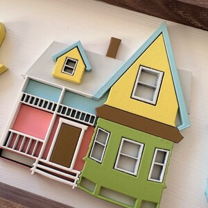 Op de afbeelding: Een geel, groen en roze houten huis met een blauw dak en witte versiering. Het huis heeft een veranda met een bruine deur en een witte railing. Er zijn ramen aan de voor- en zijkant van het huis.