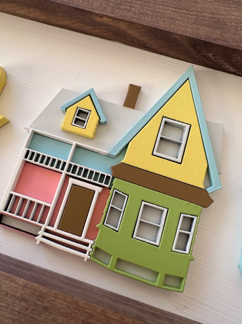 Op de afbeelding: Een geel, groen en roze houten huis met een blauw dak en witte versiering. Het huis heeft een veranda met een bruine deur en een witte railing. Er zijn ramen aan de voor- en zijkant van het huis.