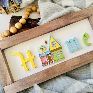 Op de afbeelding: Een houten bord met het woord "Home" in reli&euml;f, gestileerde letters. De "H" is geel, de "o" is blauw, de "m" is blauw en de "e" is groen. Een klein, geel, blauw en groen huis is gecentreerd op het bord.
