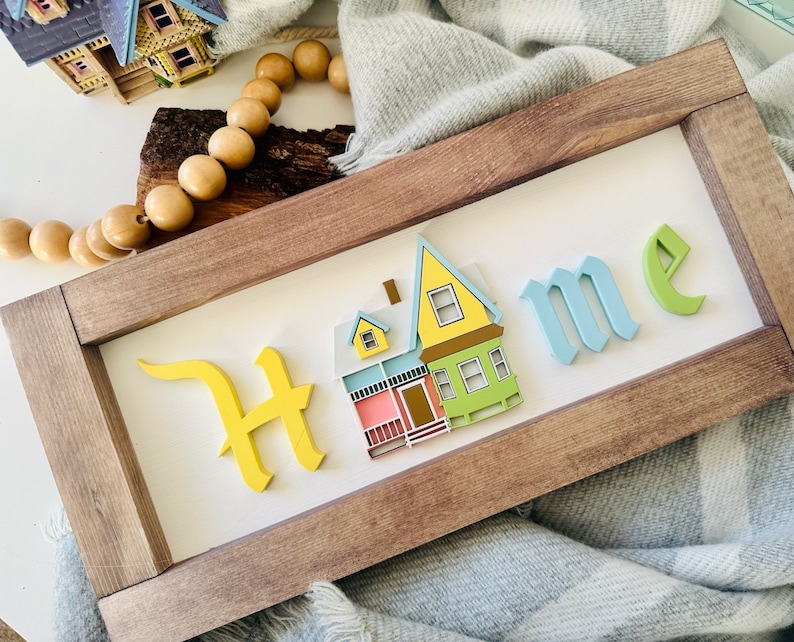 Op de afbeelding: Een houten bord met het woord "Home" in reli&euml;f, gestileerde letters. De "H" is geel, de "o" is blauw, de "m" is blauw en de "e" is groen. Een klein, geel, blauw en groen huis is gecentreerd op het bord.