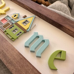 Op de afbeelding: Een houten bord met het woord "home" gespeld in 3D-letters. De letters zijn in verschillende kleuren geverfd, waaronder blauw, groen en geel. Het bord heeft ook een klein, 3D-model van een huis, geverfd in geel, groen en roze.