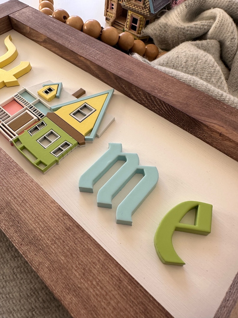 Op de afbeelding: Een houten bord met het woord "home" gespeld in 3D-letters. De letters zijn in verschillende kleuren geverfd, waaronder blauw, groen en geel. Het bord heeft ook een klein, 3D-model van een huis, geverfd in geel, groen en roze.