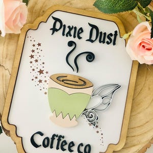 Könnte beinhalten: Holzschild mit den Worten "Pixie Dust Coffee co" in schwarzer Schrift. Das Schild zeigt ein Kaffeetassen-Design mit einem grün-beigen Farbschema und einem dekorativen flügelförmigen Griff. Das Schild befindet sich auf einem Holzgrund.