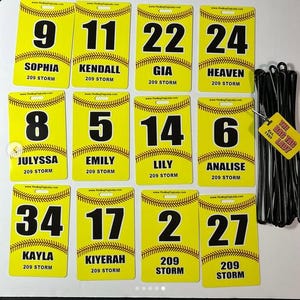 May include: A collection of yellow bag tags with black numbers and names, including "Sophia," "Kendall," and "Gia." Each tag features a baseball stitch design and the text "209 STORM." A tag with the text "THE BAG TAG LADY Est. 1995" is also visible.