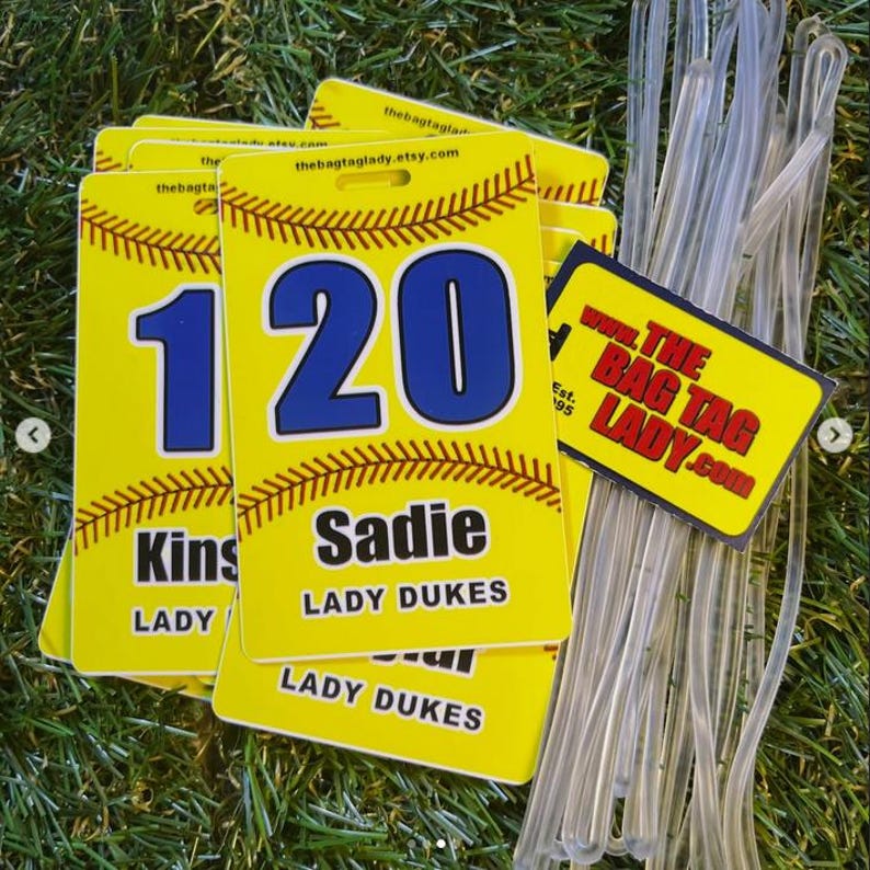May include: Yellow bag tags with baseball stitching, featuring the names "Kins" and "Sadie", and the number "120" in blue. The tags also display "LADY DUKES" and the website "thebagtaglady.etsy.com". Clear plastic loops are included.