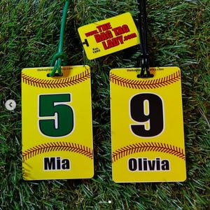 May include: Two yellow luggage tags with baseball stitching and the text "Mia" and "Olivia" below large numbers 5 and 9. A smaller tag reads "THE BAG TAG LADY.com" and "Est. 1995". The tags hang on green and black cords.