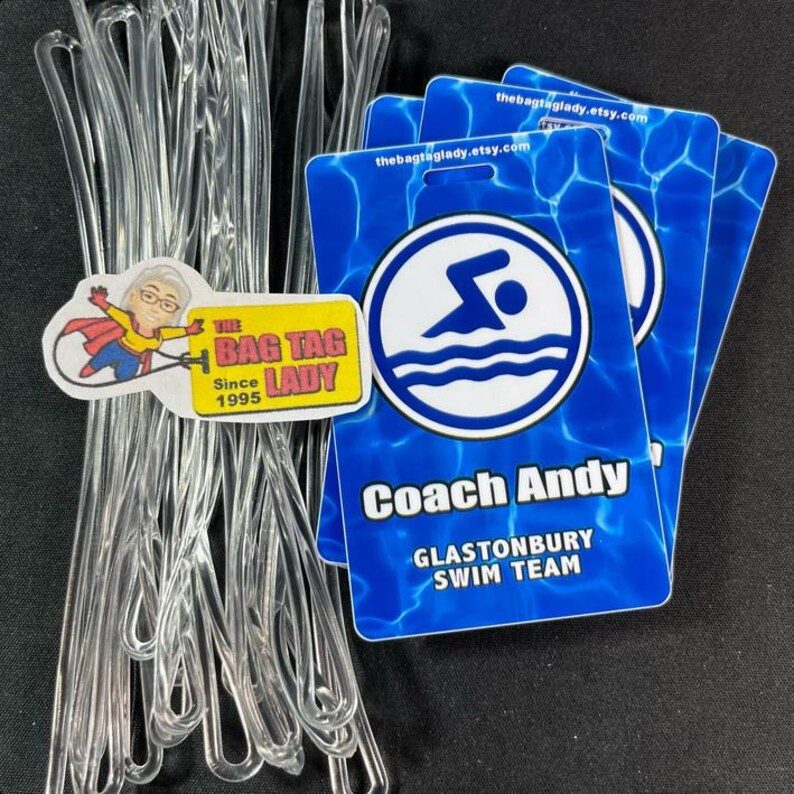 May include: Clear plastic bag tag loops and blue swim team tags. The tags have a swimmer graphic and read "Coach Andy" and "Glastonbury Swim Team". A sticker with "The Bag Tag Lady Since 1995" is also present.