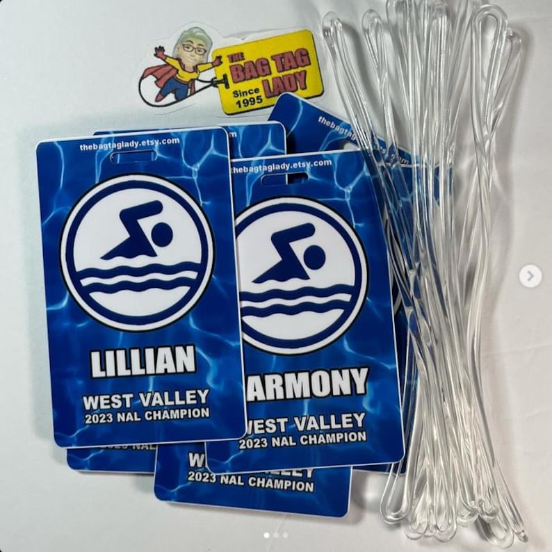 May include: Blue and white swimming-themed bag tags with the text "LILLIAN" and "ARMONY", along with "WEST VALLEY 2023 NAL CHAMPION". The tags feature a swimmer graphic and the website thebagtaglady.etsy.com. Clear plastic loops are visible.