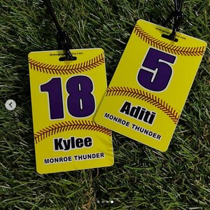 May include: Two yellow softball bag tags with black lanyards. One tag displays the number 18 in purple, the name Kylee, and "Monroe Thunder." The other tag shows the number 5, the name Aditi, and "Monroe Thunder." Both tags have baseball stitching.