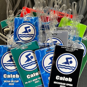 May include: A collection of colorful luggage tags with clear plastic loops. Each tag features a swimmer graphic within a white circle, with the text "Caleb" and event details like "MCSA JR CUP 2020" and "MICHIGAN OPEN 2022".
