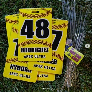 May include: Yellow softball bag tags with black numbers and text. The tags feature a baseball stitch design and the text "RODRIGUEZ" and "APEX ULTRA". The tags are on a green surface.