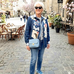May include: A woman in a denim jacket adorned with floral and lace details, blue jeans, and a light blue crossbody bag. She is wearing sunglasses and trainers. The background features a European street with cafes and blossoming trees.