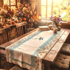 Op de afbeelding: Een rechthoekige beige tafelloper met een centraal geborduurd bladmotief, geflankeerd door ingewikkelde blauwgroene en crèmekleurige kanten randen. De loper ligt op een houten tafel en heeft een textielafwerking met een duidelijke textuur.