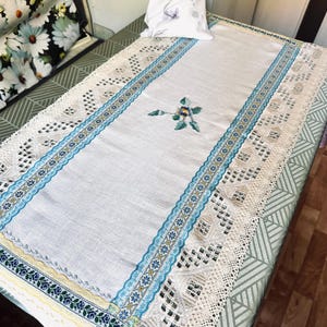 Op de afbeelding: Een rechthoekige witte linnen tafelloper met een centraal geborduurd bloemmotief in blauw, geel en groen. De loper is afgezet met ingewikkeld crèmekleurig kant, een blauw lint met patroon en een dunne gele rand met blauwe bloemaccenten.
