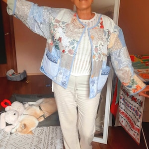May include: A patchwork jacket with a round collar and light blue trim. The jacket features various fabric patterns in shades of blue, beige, and floral prints. The person is wearing a white top and beige pants. The jacket has two pockets.