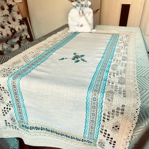 Op de afbeelding: Een rechthoekige tafelloper van lichtgekleurde stof met een centraal geborduurd bloemmotief in blauw en groen. De loper is afgezet met ingewikkelde turquoise en witte kanten randen met geometrische patronen en geschulpte randen.