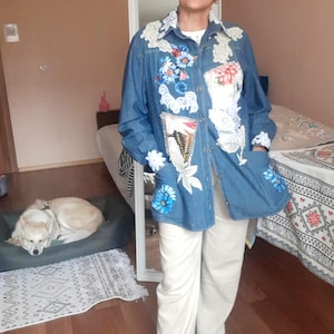 May include: A denim jacket with floral and butterfly embroidery, paired with white pants and sneakers. The jacket features a variety of colorful flower designs and lace trim. The person is wearing sunglasses.