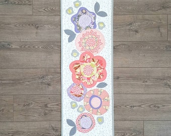 Puede incluir: Un camino de mesa blanco con un borde gris presenta una fila vertical de cinco flores de tela de colores. Las flores son rosas, moradas y amarillas sobre un fondo blanco. Las flores están dispuestas en un patrón escalonado.