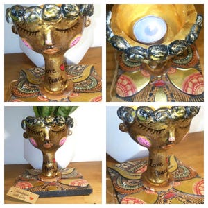 May include: A handcrafted, gold-toned candle holder with a face design, featuring closed eyes and a serene expression. The base is adorned with a decorative pattern. The words "Love Peace Hope" are written on the stem. A small heart is also present.
